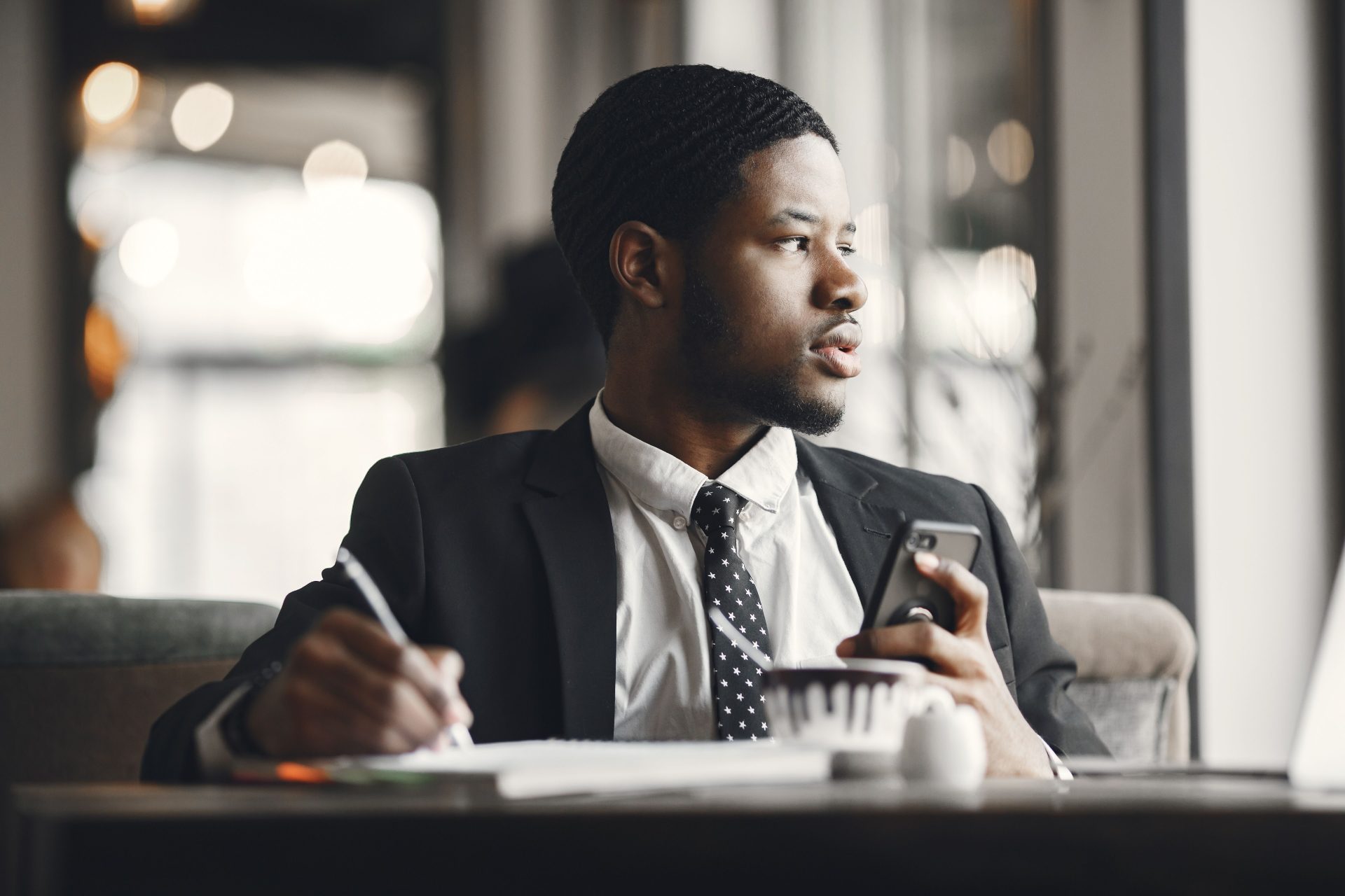 Young businessman in formal attire using mobile phone and writing at desk in office